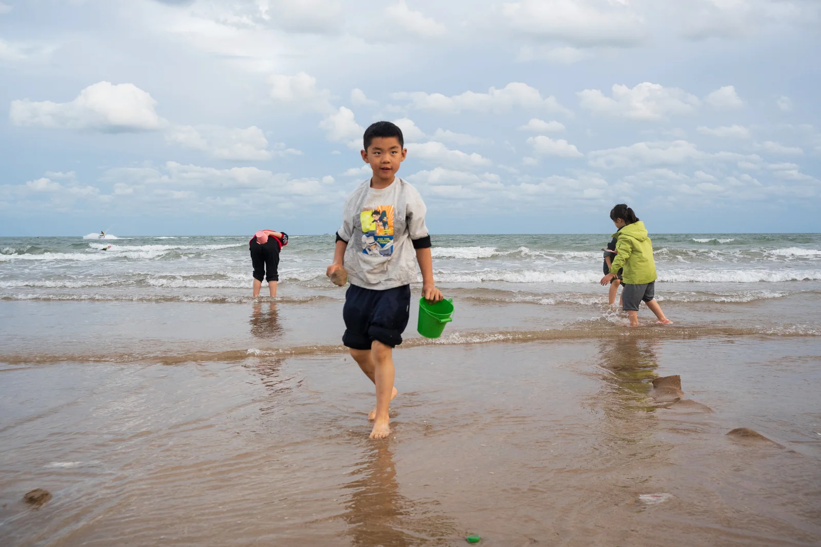 男孩提着绿色水桶从浅水区走来，身后游客在海边玩耍